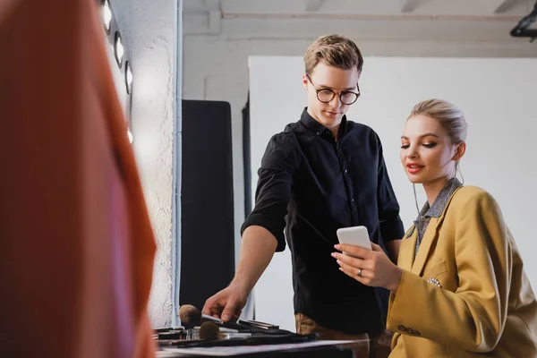 Makeup Artist taking cosmetic brush and model looking at smartphone on backstage — Stock Photo