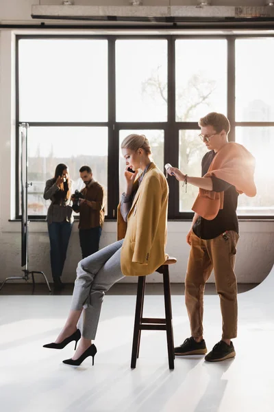 Stylist using lint roller, model talking on smartphone, producer and photographer talking on backstage — Stock Photo