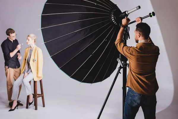 Foco seletivo do fotógrafo tocando refletor nos bastidores e maquiador fazendo maquiagem para modelar — Fotografia de Stock