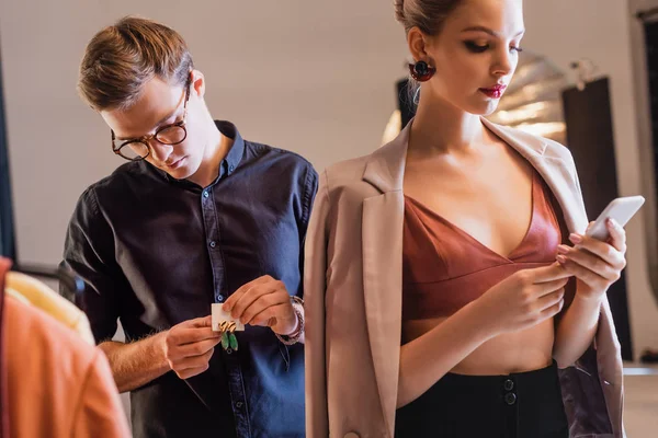 Orecchini stylist holding e modello elegante utilizzando smartphone sul backstage — Foto stock
