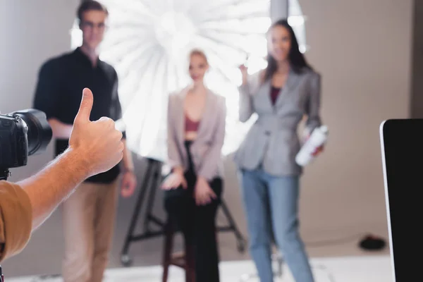 Selective focus of photographer showing thumb up to model, stylist, hairstylist on backstage — Stock Photo