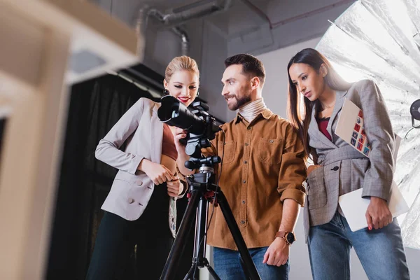 Vue en angle bas du photographe, modèle et producteur souriant regardant l'appareil photo numérique dans les coulisses — Photo de stock