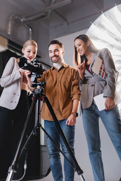 Visão de baixo ângulo do fotógrafo sorridente, modelo e produtor olhando para a câmera digital nos bastidores — Fotografia de Stock