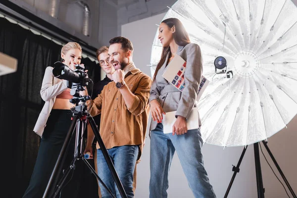 Vista de bajo ángulo del fotógrafo sonriente, modelo, estilista y productor mirando a la cámara digital en el backstage - foto de stock