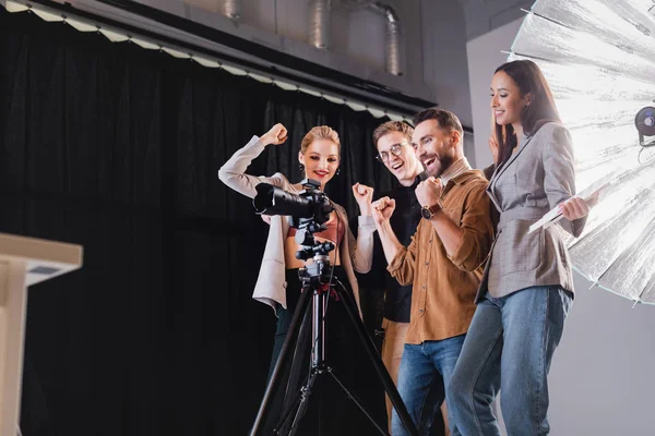 Vista de bajo ángulo de fotógrafo sonriente, modelo, estilista y productor mirando a la cámara digital y mostrando sí gesto en el backstage - foto de stock