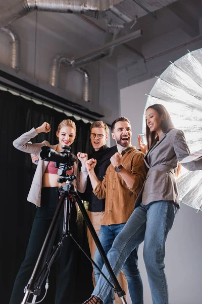 Vista de bajo ángulo de fotógrafo sonriente, modelo, estilista y productor mirando a la cámara digital y mostrando sí gesto en el backstage - foto de stock