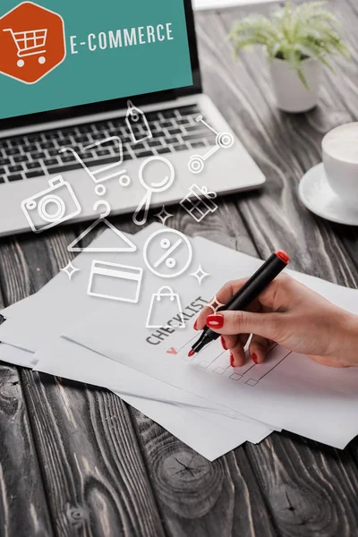 Vista ritagliata di donna d'affari in possesso di pennarello rosso vicino checklist, illustrazione, tazza e laptop con lettere di e-commerce — Foto stock