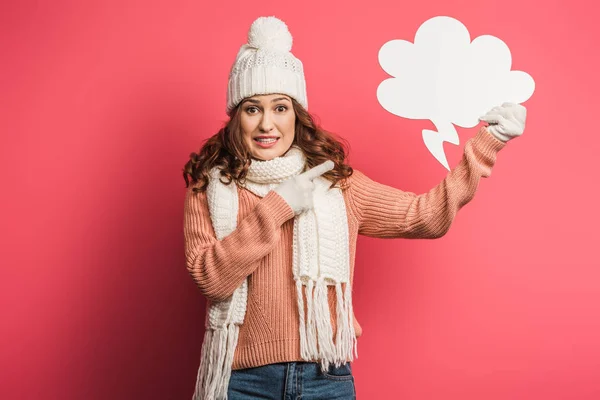 Chica impactada en sombrero caliente y bufanda señalando con el dedo en la burbuja de pensamiento sobre fondo rosa - foto de stock