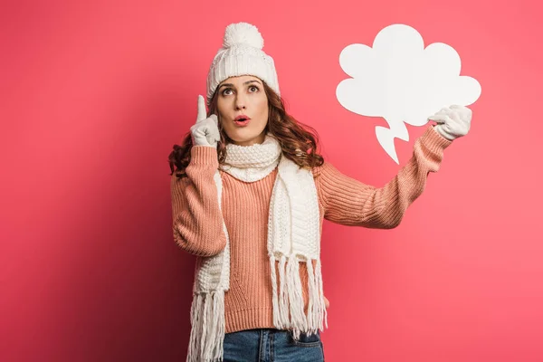 Chica reflexiva en sombrero caliente y bufanda que muestra signo de idea mientras sostiene la burbuja de pensamiento sobre fondo rosa - foto de stock