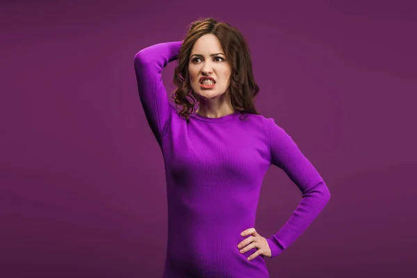 Mujer enojada de pie con la mano en la cadera y tocando la cabeza sobre fondo púrpura - foto de stock