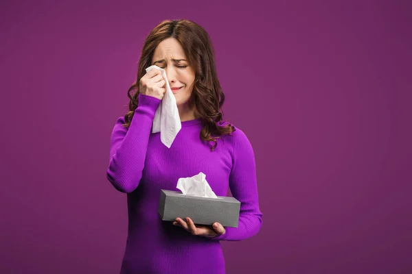Femme bouleversée essuyant des larmes avec une serviette en papier tout en pleurant sur fond violet — Photo de stock