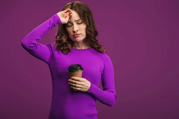 Chica agotada tocando la frente mientras sostiene el café para ir sobre fondo púrpura - foto de stock