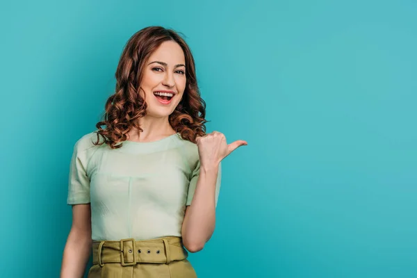 Smiling woman pointing with thumb and looking at camera on blue background — Stock Photo