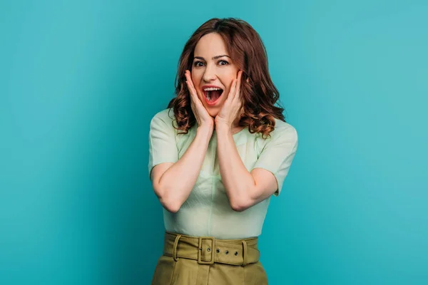 Femme choquée toucher le visage tout en se tenant debout avec la bouche ouverte sur fond bleu — Photo de stock