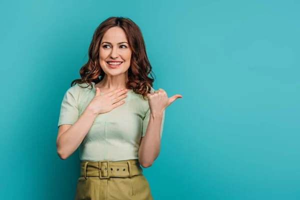 Mujer sorprendida y sonriente señalando con el pulgar y sosteniendo la mano en el pecho sobre fondo azul - foto de stock