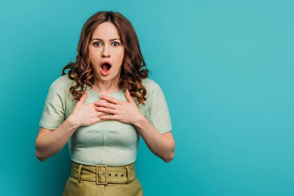 Schockierte Frau hält Hände auf Brust, während sie mit offenem Mund vor blauem Hintergrund steht — Stockfoto