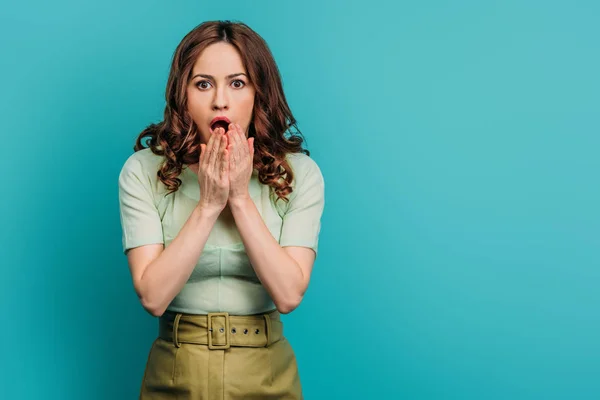 Femme choquée tenant les mains près du visage tout en se tenant la bouche ouverte sur fond bleu — Photo de stock