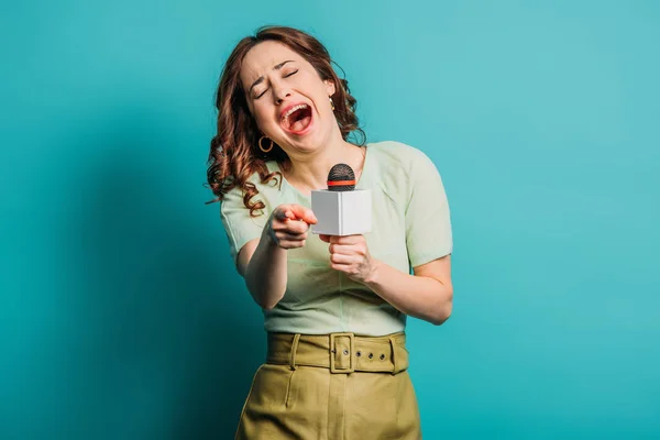 Cheerful journalist pointing with finger at camera while holding microphone on blue background — Stock Photo