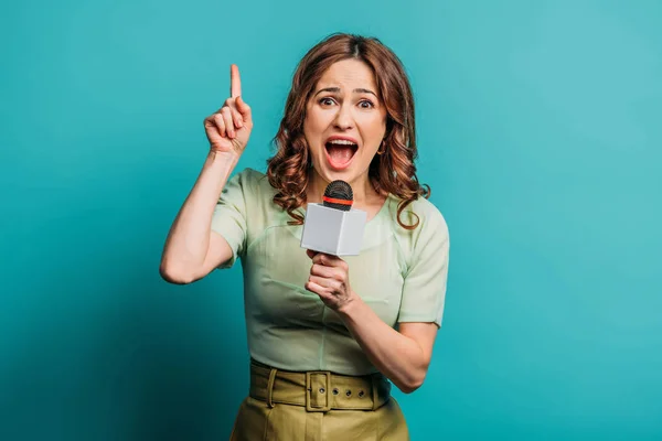Angry journalist pointing up with finger while shouting in microphone on blue background — Stock Photo