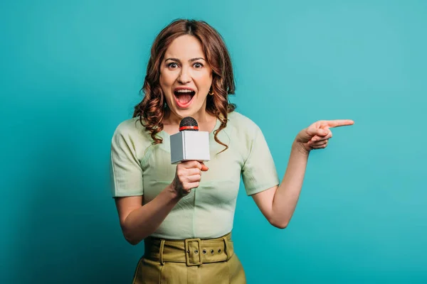 Excited journalist pointing with finger while holding microphone on blue background — Stock Photo