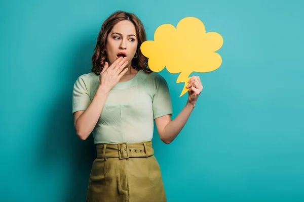 Schockierte Frau hält Hand in Mund, während sie Gedankenblase auf blauem Hintergrund hält — Stockfoto