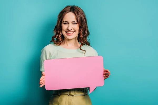 Cheerful woman looking at camera while holding speech bubble on blue background — Stock Photo