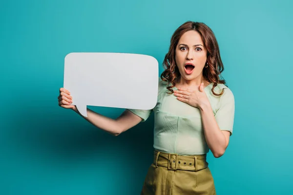 Shocked woman touching chest while holding speech bubble on blue background — Stock Photo