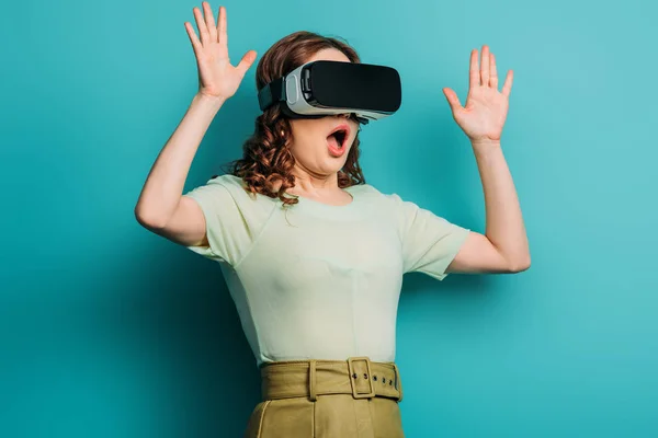 Chica asustada en auriculares vr de pie con las manos levantadas sobre fondo azul - foto de stock