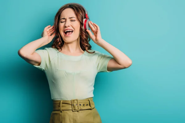 Alegre chica cantando en auriculares inalámbricos con los ojos cerrados sobre fondo azul - foto de stock