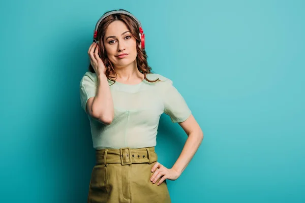 Chica escéptica en auriculares inalámbricos de pie con la mano en la cadera sobre fondo azul - foto de stock