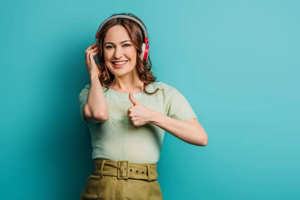 Menina feliz em fones de ouvido sem fio mostrando polegar no fundo azul — Fotografia de Stock