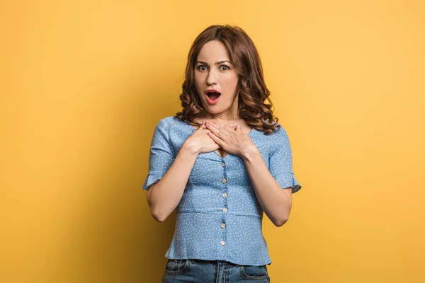 Surprised woman holding hands on chest while looking at camera on yellow background — Stock Photo