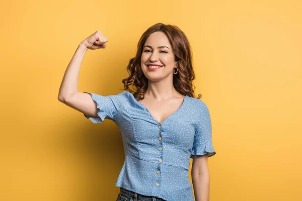 Fröhliche Frau, die Macht zeigt, während sie in die Kamera auf gelbem Hintergrund schaut — Stockfoto