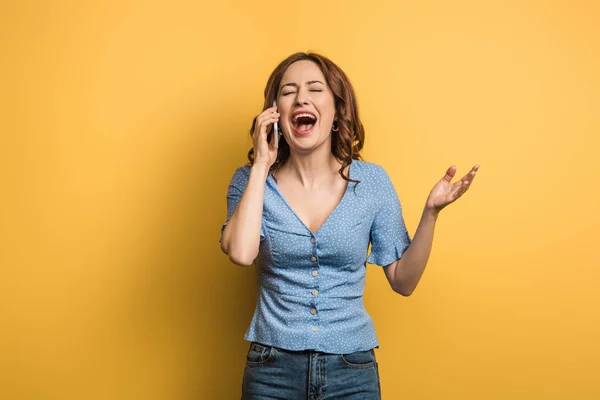 Mujer alegre riendo con los ojos cerrados mientras habla en el teléfono inteligente sobre fondo amarillo - foto de stock