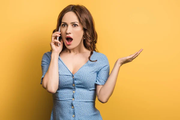 Mujer sorprendida de pie con el brazo abierto mientras habla en el teléfono inteligente sobre fondo amarillo - foto de stock