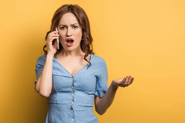 Mujer enojada de pie con el brazo abierto mientras habla en el teléfono inteligente sobre fondo amarillo - foto de stock