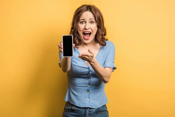 Femme choquée pointant du doigt le smartphone avec écran blanc sur fond jaune — Photo de stock