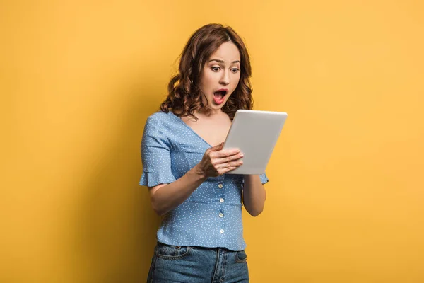 Mulher chocada usando tablet digital no fundo amarelo — Fotografia de Stock