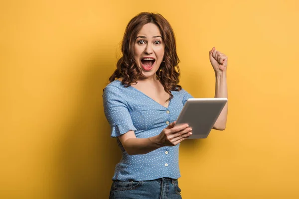 Aufgeregte Frau zeigt Siegergeste, während sie ein digitales Tablet auf gelbem Hintergrund hält — Stockfoto