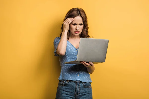 Thoughtful girl holding hand near head while looking at laptop on yellow background — Stock Photo