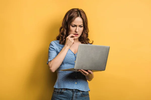 Thoughtful girl holding hand near face while looking at laptop on yellow background — Stock Photo