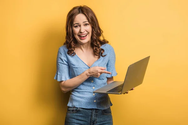 Cheerful girl looking at camera while pointing with finger at laptop on yellow background — Stock Photo
