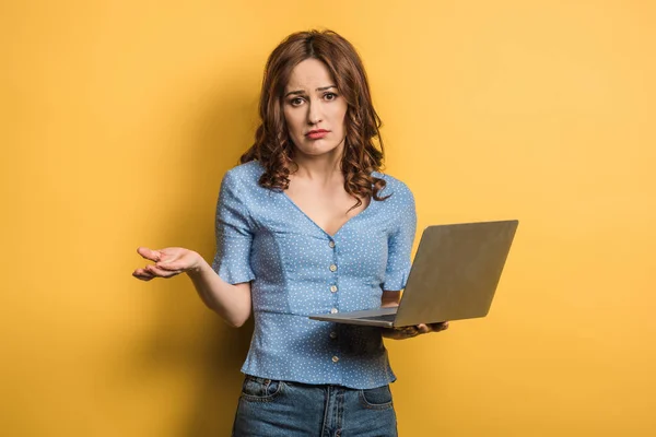Chica confundida mostrando gesto encogiéndose de hombros mientras sostiene el ordenador portátil sobre fondo amarillo — Stock Photo
