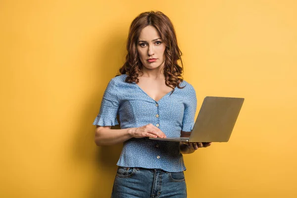 Displeased girl looking at camera while holding laptop on yellow background — Stock Photo