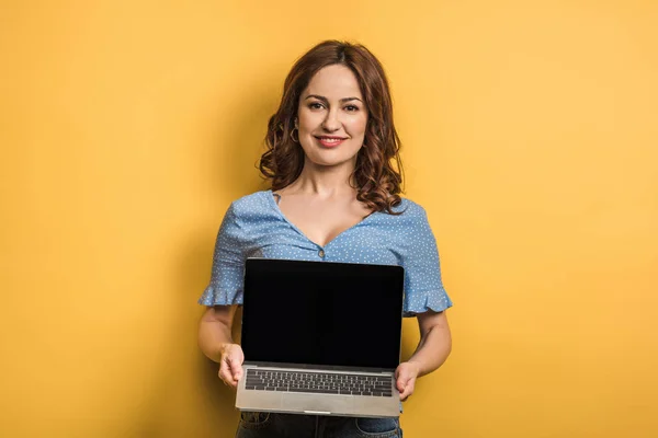 Усміхнена жінка тримає ноутбук з порожнім екраном на жовтому тлі — стокове фото