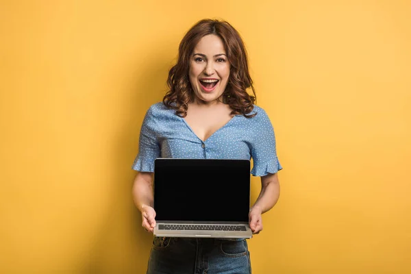 Весела жінка сміється, тримаючи ноутбук з порожнім екраном на жовтому тлі — стокове фото