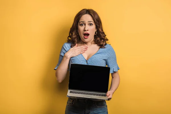 Donna scioccata toccando petto tenendo il computer portatile con schermo bianco su sfondo giallo — Foto stock