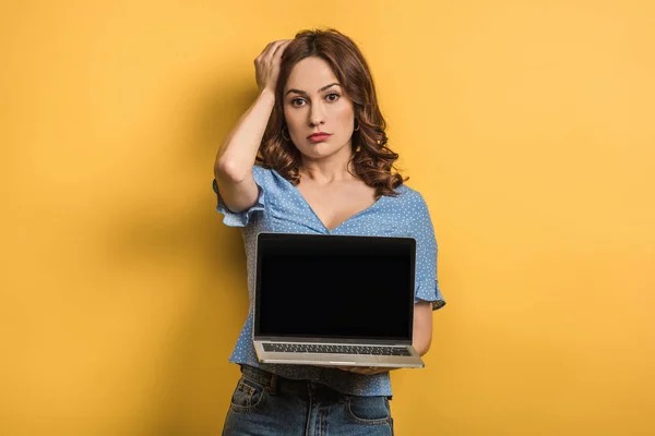 Mujer disgustada tocando la cabeza mientras sostiene el ordenador portátil con pantalla en blanco sobre fondo amarillo - foto de stock