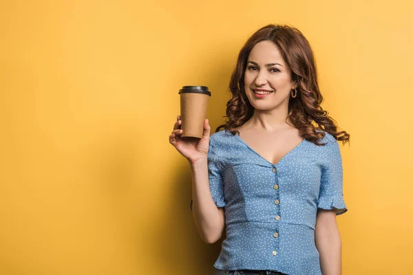 Smiling woman holding coffee to go and looking at camera on yellow background — Stock Photo