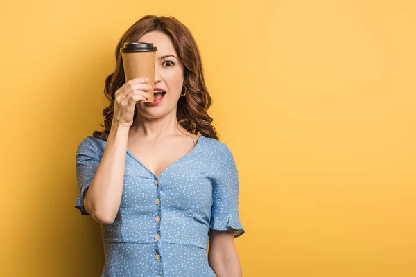 Gaie femme couvrant oeil avec tasse en papier sur fond jaune — Photo de stock